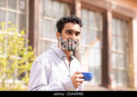 A smiling man with a beard holds a blue cup outdoors, standing in a sunny area Stock Photo