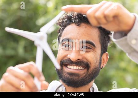 A smiling man with curly hair holds a small white wind turbine model above his head against a blurred green foliage background. Stock Photo