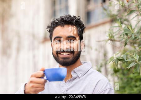 A smiling man with curly hair holds a blue cup towards the camera, standing in front of a blurred background Stock Photo