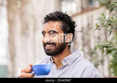 A smiling man with dark curly hair, holding a blue cup, enjoying a moment outdoors surrounded by green Stock Photo