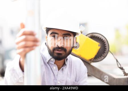 A construction worker in a white hard hat and shirt is gripping a metallic pole with yellow construction equipment in the background. Stock Photo