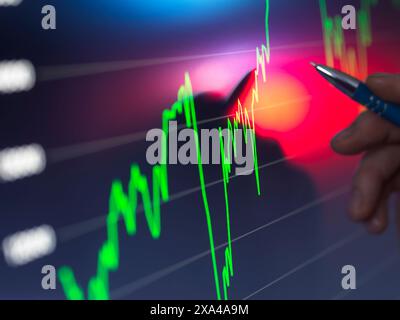 A hand holding a pen points to a bright stock market chart on a digital screen with fluctuating green and red graph lines indicating financial data. Stock Photo