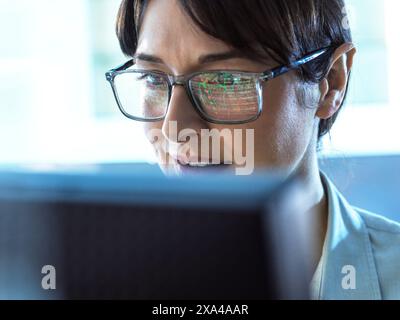 A professional woman is intensely focused on the content displayed on her computer monitor, which is reflected in her glasses. Stock Photo