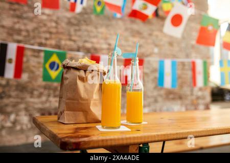 A table with two bottles of orange juice and a brown paper takeout bag against a backdrop of international flags. Stock Photo