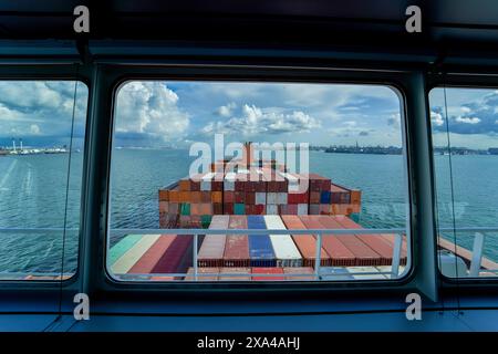 A view from a ship's bridge window overlooking a large stack of colorful shipping containers on the vessel's deck, with the ocean and cloudy sky in the background. Stock Photo