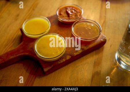 A wooden serving board with three small bowls of sauce and a jar of sauce on a wooden table. Stock Photo