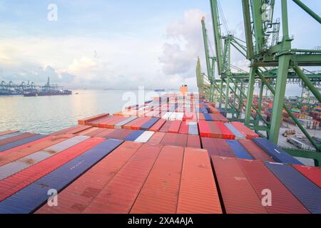 A container ship is docked at a busy port with a clear sky at dawn or dusk, as container cranes extend over the colorful array of shipping containers. Stock Photo
