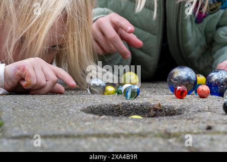 A close-up view of a child's hands playing marbles on a concrete ground, with colorful glass marbles scattered around a small hole. Stock Photo