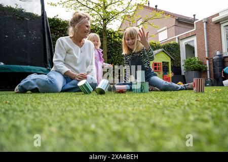An elderly woman and two young girls are sitting on a grass lawn playing with colorful tin cans, set up like bowling pins, with a child's playhouse in the background. Stock Photo