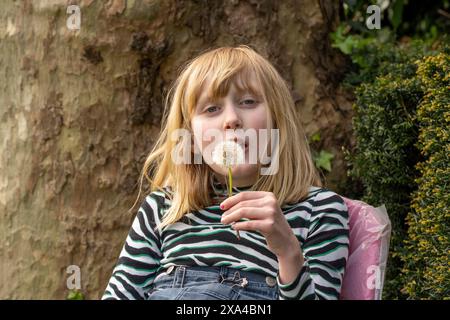 A young girl with blonde hair is sitting outdoors, blowing on a dandelion seed head in a relaxed setting, with trees and shrubbery in the background. Stock Photo