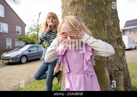 A young girl in a pink dress is covering her eyes with her hands while standing in front of a tree, as a woman playfully acts shocked in the background on a residential street. Stock Photo