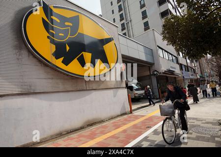 A woman rides a bicycle past a large logo for Yamato Delivery Company on a depot in Ginza, Tokyo, Japan. Stock Photo