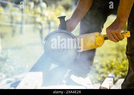 Person using big yellow grinder to cut concrete poles in home garden. Spinning metal diamond disc to cut concrete, visible rotating blade. Dust and de Stock Photo