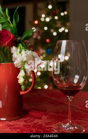 A festive table setting featuring a bouquet of flowers in a red vase and an empty wine glass, with a blurred Christmas tree adorned with lights in the background. Stock Photo