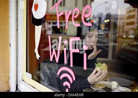 A person seen through a window sits inside a cafÈ, eating a sandwich with a Free WiFi' sign painted on the glass.' Stock Photo