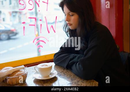 A woman sits contemplatively by a window in a cafe, with a cup of cappuccino and a sandwich on the table. Stock Photo