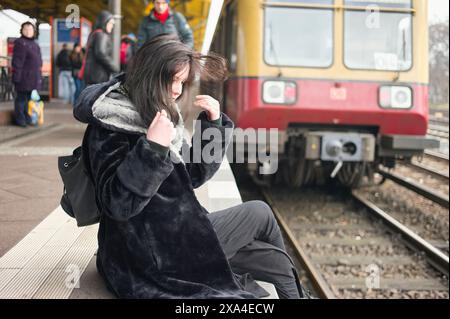 A person sits on the edge of a train platform, with hair obscuring their face, as a train arrives in the background. Stock Photo