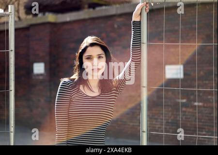 A woman with long hair poses confidently with her arm raised, holding onto a metal fence, with sunlight casting a warm glow behind her. Stock Photo