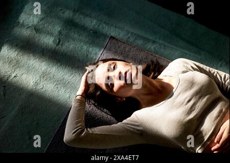 A woman is lying down on a dark surface with her head resting on her hand, bathed in a shaft of sunlight. Stock Photo
