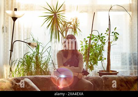 A young woman sits on a couch surrounded by potted plants in a warmly lit room, with sunlight filtering through the window behind her. Stock Photo