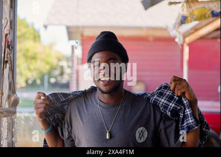 A smiling young man with a beanie stands outside, holding the collars of his plaid shirt with sunlit foliage in the background. Stock Photo