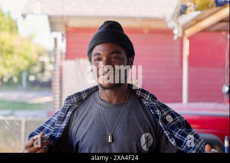 A smiling man wearing a beanie and plaid shirt stands in front of a house with a red door. Stock Photo