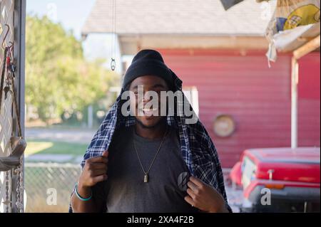 A smiling young man wearing a beanie and hooded plaid shirt stands outside with a red building and a vehicle in the background. Stock Photo