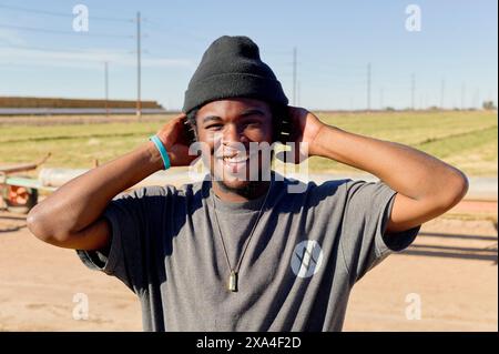 A smiling young man stands outdoors wearing a black beanie and t-shirt, with a sunny blue sky and open field in the background. Stock Photo