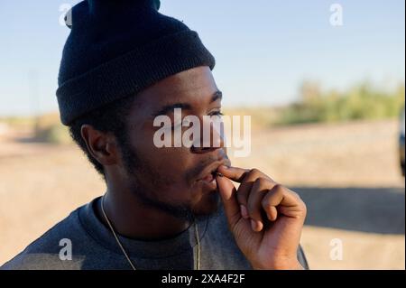 A young man wearing a beanie is outdoors, partially lit by sunlight, looking to the side while thoughtfully biting his fingernail. Stock Photo