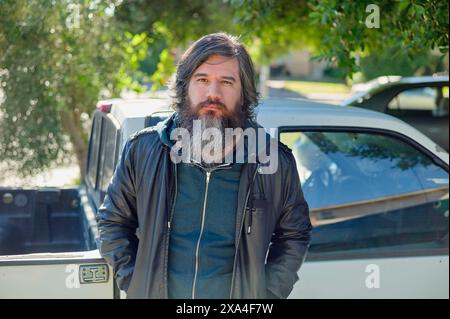 A bearded man stands in front of a white pickup truck, with greenery and a residential setting in the background. Stock Photo
