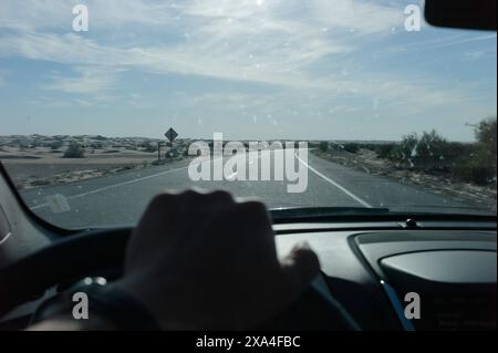 A first-person view from inside a vehicle showing a hand on the steering wheel, driving on an open road through a sandy desert landscape with a clear blue sky overhead. Stock Photo