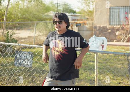 Man in sunglasses and black t-shirt leaning on a chain-link fence with Beware of Dog' sign.' Stock Photo