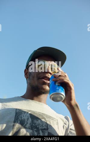 A man wearing sunglasses and a cap enjoys a beverage from a can against a clear sky. Stock Photo