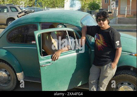 Two young men are standing next to a vintage green Volkswagen Beetle, one sitting inside with the door open and the other leaning on the door from outside. Stock Photo
