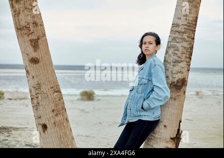 A woman stands between two palm trees on a cloudy day, her arms crossed as she gazes into the distance with a thoughtful expression. Stock Photo