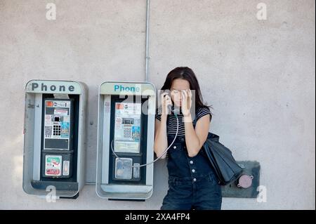 A woman is standing between two public payphones, engaged in a conversation on one of them, with her eyes closed and a slight frown, possibly indicating a serious or troubling discussion. Stock Photo