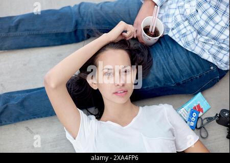 A young woman is lying on the floor next to a man, holding her hair up and looking directly at the camera, with a relaxed expression. Stock Photo