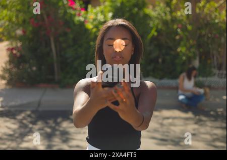 A focused young woman holds a camera lens up to her eye, taking a photo, with another person blurred in the background sitting on a bench surrounded by green foliage. Stock Photo