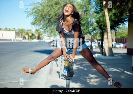 A young woman in a black sheer top and shorts confidently poses on a city sidewalk with one foot on a parking meter, laughing and looking upwards. Stock Photo