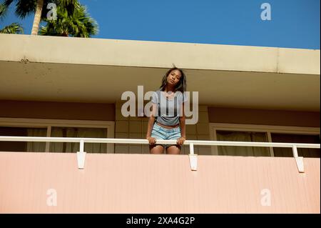 A smiling young woman leans on a balcony railing, with a blue sky and palm trees in the background. Stock Photo