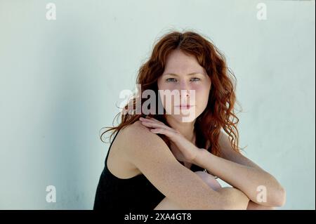 A red-haired woman with a subtle gaze sits against a white wall, her head resting on her hand. Stock Photo