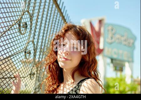 A smiling woman with red hair is leaning against a chain-link fence, with a vintage-looking 'Boulder Hotel' sign in the blurred background. Stock Photo