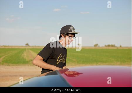 A smiling young man wearing a black cap and glasses stands beside a red car with a rural landscape in the background. Stock Photo