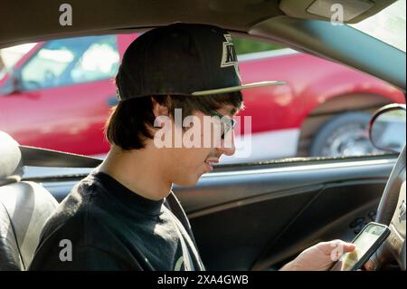 A young man wearing glasses and a baseball cap is sitting in the driver's seat of a car, smiling as he looks down at a smartphone in his hands. Stock Photo