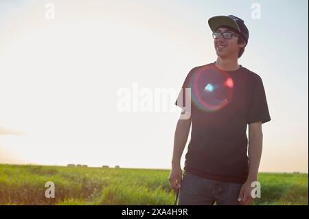 A smiling young man wearing glasses, a cap, and a black t-shirt stands in a field with the sun setting in the background, casting a warm glow. Stock Photo
