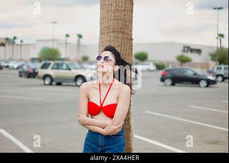 A woman in a red bikini top and blue jeans leans against a palm tree in a parking lot, looking upwards with her hair blowing in the wind. Stock Photo