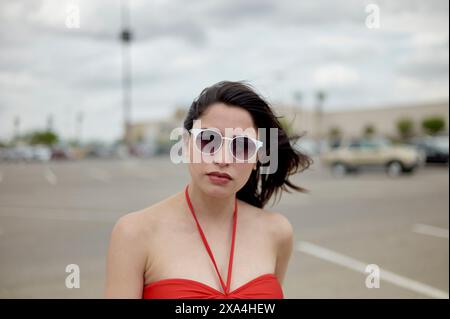 A woman wearing sunglasses and a red dress stands in a parking lot with a cloudy sky overhead and a classic car in the background. Stock Photo