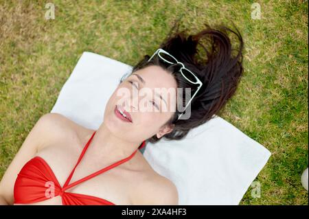 A woman is lying on her back on a white towel placed on grass, wearing a red swimsuit and sunglasses on her head, with a relaxed smile on her face. Stock Photo