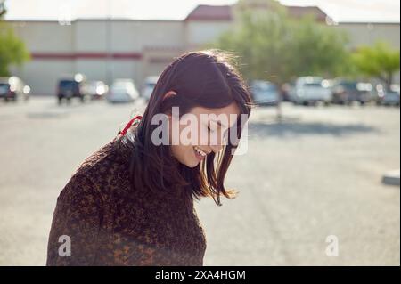 A smiling young woman with dark hair enjoying a sunny day in a parking lot, with buildings visible in the background. Stock Photo