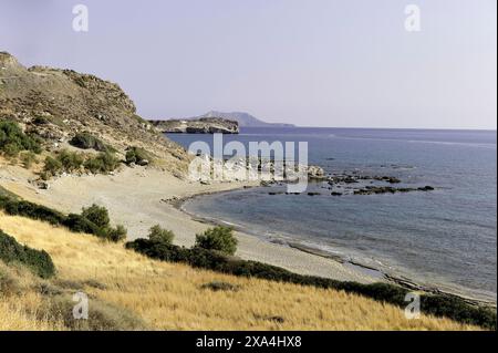 A serene view of a tranquil beach with a rocky hillside, golden dry grass in the foreground, and calm blue waters extending to the horizon under a clear sky. Stock Photo